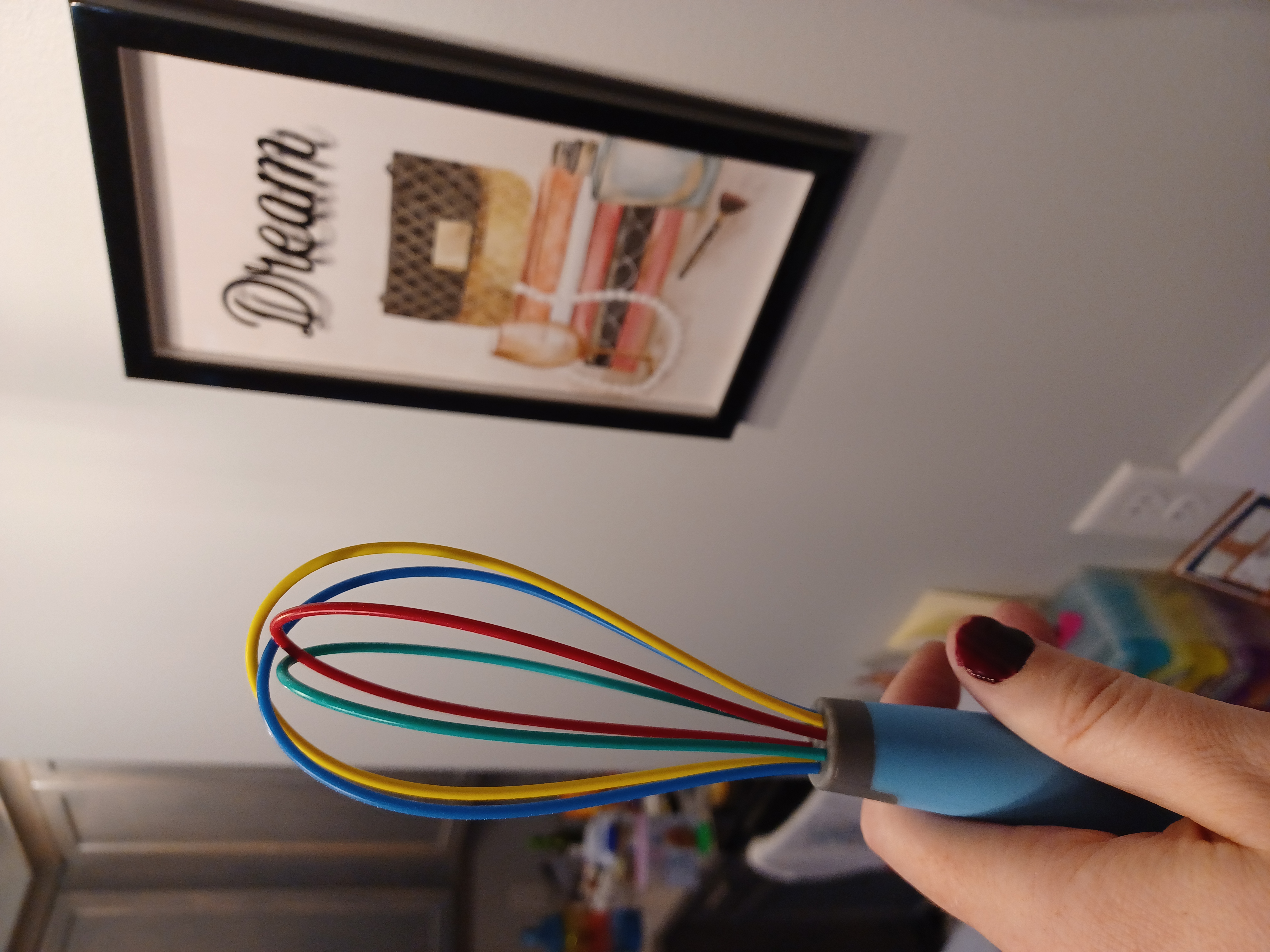 a rainbow colored whisk held up by a hand with painted fingernails but shaped by testosterone-driven puberty with a framed print in the background with the word dream in cursive font and a stack of books a perfume bottle and a necklace